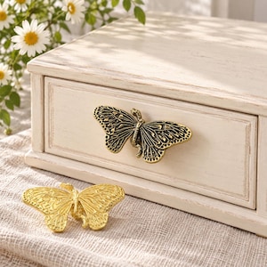 May include: A cream-colored wooden drawer unit featuring a dark metallic butterfly-shaped handle. A second, bright gold-toned butterfly-shaped handle rests on the textured fabric surface in front of the drawer unit.