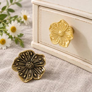 May include: Two flower-shaped cabinet knobs made of metal. One knob is bright gold and attached to a white wooden drawer, while the other is an antique brass finish and rests on a textured fabric surface. Both feature detailed petals.