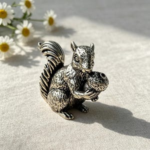 May include: A small, metallic silver-toned squirrel figurine with a textured fur finish and a bushy tail. The squirrel is depicted in a seated position, holding a textured acorn in its front paws. The surface of the metal shows dark oxidation.