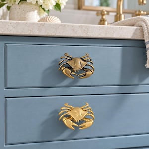 Puede incluir: Dos pomos de metal para armarios con forma de cangrejo fijados en un frente de cajón azul. El pomo superior tiene un acabado en bronce antiguo, mientras que el inferior presenta un acabado dorado brillante y pulido.
