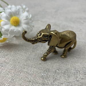 May include: A small, brass-coloured elephant figurine with detailed trunk, ears, and legs. The elephant is walking, with its trunk raised. The figurine is on a textured, neutral-coloured surface. Daisies are in the background.