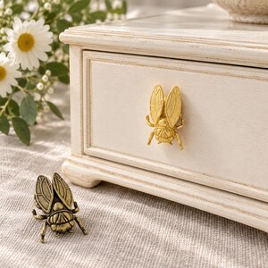May include: A gold-toned bee-shaped drawer knob attached to a white wooden drawer front, with a matching antique brass-toned bee-shaped knob resting on the surface below. The drawer features a rectangular frame molding detail.