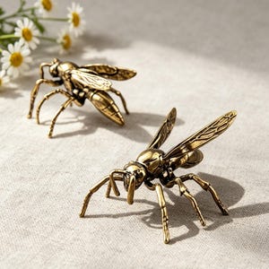May include: Two metallic wasp figurines crafted with a polished antique brass finish. Each insect sculpture features detailed wings with etched vein patterns, segmented bodies, and six spindly legs positioned to stand upright on a flat surface.