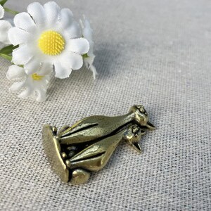 Op de afbeelding: Een bronzen metalen bedel met twee gestileerde vogels. De bedel heeft een rechthoekige basis en gedetailleerde vogelkenmerken. Witte madeliefjes met gele centra bevinden zich op de achtergrond.