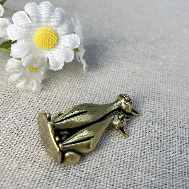 Op de afbeelding: Een bronzen metalen bedel met twee gestileerde vogels. De bedel heeft een rechthoekige basis en gedetailleerde vogelkenmerken. Witte madeliefjes met gele centra bevinden zich op de achtergrond.