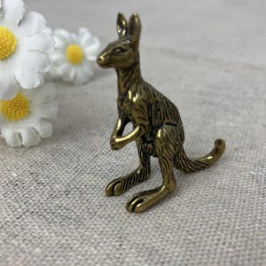 May include: A small, detailed brass kangaroo figurine. The kangaroo is posed upright with its arms folded, and its tail curled. The figurine has a textured surface and is set against a neutral background with white daisy flowers.