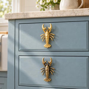 Op de afbeelding: Twee ladeknoppen in de vorm van kreeften bevestigd aan een blauwe kast. De bovenste knop heeft een gepolijste gouden afwerking, terwijl de onderste knop een verouderde, donkere messing afwerking heeft. Beide kreeften tonen gedetailleerde scharen.