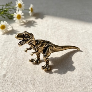 May include: A small, metallic, brass-colored Tyrannosaurus Rex figurine with a textured, antique finish. The dinosaur is depicted in a standing position with its mouth open, showing teeth, and its tail extended horizontally behind it.