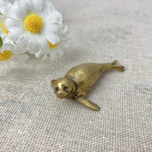 Könnte beinhalten: Kleine Robbenfigur aus Messing. Die Robbe liegt mit ausgestreckten Flossen. Die Figur hat eine glatte, polierte Oberfläche und detaillierte Merkmale. Im Hintergrund befinden sich weiße Blumen mit gelben Zentren.