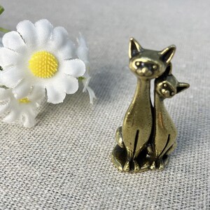 Op de afbeelding: Een kleine, bronzen kattenfiguur. De sculptuur toont twee katten, de ene iets achter de andere, in zittende positie. De katten hebben puntige oren en gedetailleerde gezichtskenmerken. Een witte madelief met een gele kern staat op de achtergrond.