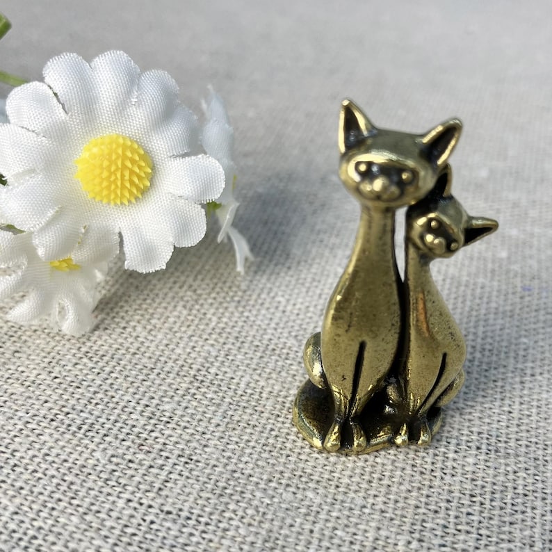 Op de afbeelding: Een kleine, bronzen kattenfiguur. De sculptuur toont twee katten, de ene iets achter de andere, in zittende positie. De katten hebben puntige oren en gedetailleerde gezichtskenmerken. Een witte madelief met een gele kern staat op de achtergrond.