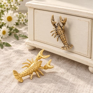 May include: Two lobster-shaped hardware pieces made of metal. One piece is finished in a dark, antique brass tone and is attached to a white wooden drawer, while the other is a bright, polished gold tone resting on a textured fabric surface.