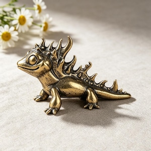 May include: A small, metallic, brass-colored lizard figurine with a textured, spiky ridge running along its back and tail. The creature has large, smooth eyes, a pointed snout, and four short legs, featuring a weathered, antique-style finish.