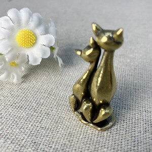 Op de afbeelding: Een kleine, bronzen kattenfiguur die twee samen zittende katten afbeeldt. De katten zijn gestileerd met langwerpige lichamen en puntige oren. Op de achtergrond een witte madelief met een gele kern.