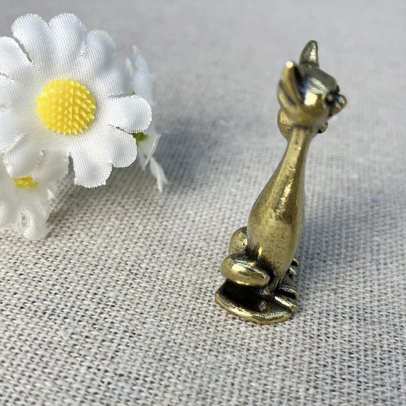 Op de afbeelding: Een kleine, antieke messing kattenfiguur in zittende positie. De kat heeft een lang, slank lichaam en puntige oren. Het beeldje is gedetailleerd met zichtbare poten en een kleine, ronde basis. Witte madeliefbloemen zijn op de achtergrond.