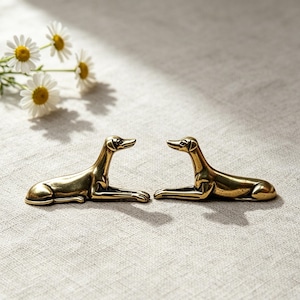 May include: Two small, polished brass figurines shaped like greyhounds in a resting, lying-down position. The metallic dog sculptures feature smooth, reflective surfaces and detailed facial features, positioned facing each other on a flat surface.