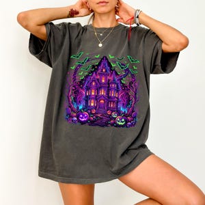 May include: A charcoal gray short-sleeve t-shirt featuring a vibrant graphic print of a haunted mansion at night. The design includes glowing purple and blue architecture, bright green bats, and orange jack-o'-lanterns set against a dark forest.