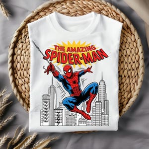 Puede incluir: Una camiseta blanca de manga corta con un gr&aacute;fico de Spider-Man con traje rojo y azul balance&aacute;ndose en una telara&ntilde;a. Sobre el personaje, el texto THE AMAZING SPIDER-MAN aparece en letras rojas sobre un sol amarillo. Debajo hay un horizonte urbano.
