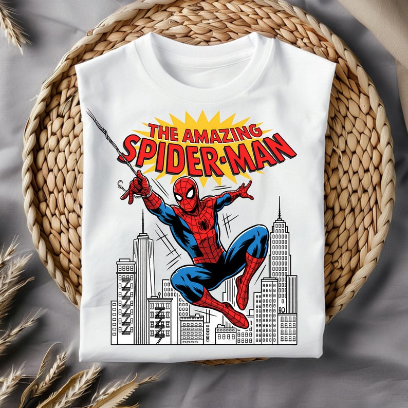 Puede incluir: Una camiseta blanca de manga corta con un gr&aacute;fico de Spider-Man con traje rojo y azul balance&aacute;ndose en una telara&ntilde;a. Sobre el personaje, el texto THE AMAZING SPIDER-MAN aparece en letras rojas sobre un sol amarillo. Debajo hay un horizonte urbano.
