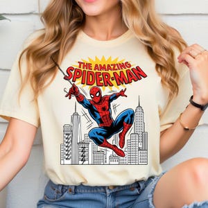 Puede incluir: Una camiseta de manga corta color crema con un estampado gr&aacute;fico de Spider-Man balance&aacute;ndose a trav&eacute;s de un horizonte urbano. El texto sobre el personaje dice THE AMAZING SPIDER-MAN en letras rojas sobre un fondo amarillo.