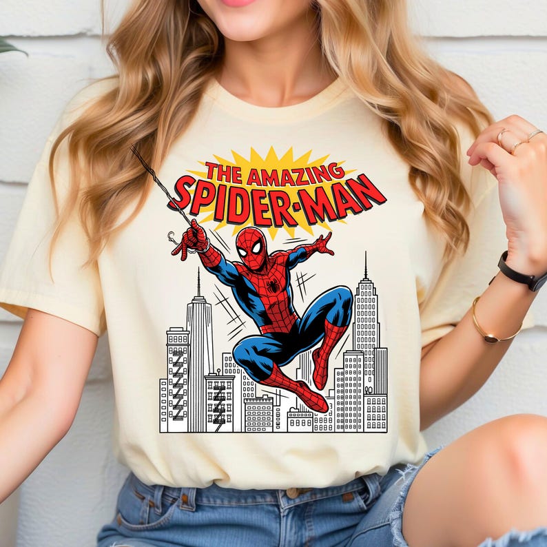 Puede incluir: Una camiseta de manga corta color crema con un estampado gr&aacute;fico de Spider-Man balance&aacute;ndose a trav&eacute;s de un horizonte urbano. El texto sobre el personaje dice THE AMAZING SPIDER-MAN en letras rojas sobre un fondo amarillo.