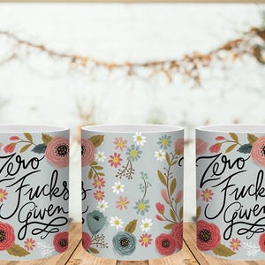 May include: Three white ceramic mugs with floral designs and the phrase "Zero Fucks Given" in black script. The mugs feature pink, red, and blue flowers on a light gray background.