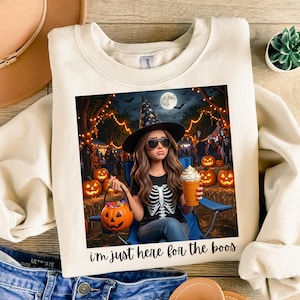 May include: A cream-coloured crewneck sweatshirt featuring a graphic print of a person wearing a witch hat, sunglasses, and a skeleton shirt, holding a pumpkin bucket and a drink. The text below reads, i'm just here for the boos.
