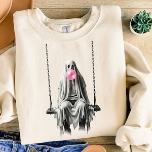 May include: A cream-colored crewneck sweatshirt featuring a graphic print of a ghost draped in a white sheet, sitting on a black chain swing and blowing a large, round, bright pink bubblegum bubble. The ghost has dark, hollow eyes.