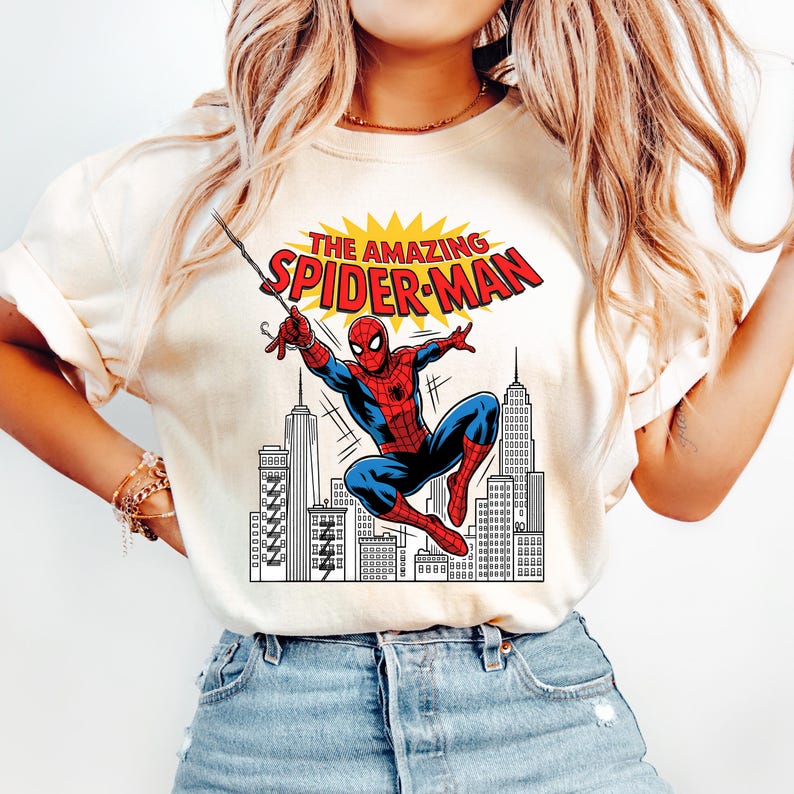 Puede incluir: Una camiseta de color crema con un estampado gr&aacute;fico de Spider-Man balance&aacute;ndose sobre el horizonte de una ciudad. El texto sobre el personaje dice THE AMAZING SPIDER-MAN en letras rojas y amarillas. El dise&ntilde;o incluye un fondo urbano en blanco y negro.