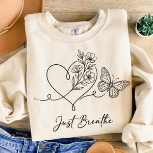 May include: A cream-colored crewneck sweatshirt featuring a black line art graphic of a heart intertwined with a floral stem and a butterfly. The text "Just Breathe" is printed in a black cursive script below the central heart and floral design.