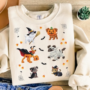 May include: A cream-colored crewneck sweatshirt featuring a graphic print of various animals in Halloween costumes, including a dog as a witch, a dog with a pumpkin head, a dog as a vampire, a dog as a mummy, and a cat with a witch hat.
