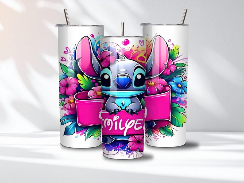 K&ouml;nnte beinhalten: Drei wei&szlig;e Edelstahlbecher mit einem Cartoon-Charakter-Design. Das Design zeigt eine blau-graue, au&szlig;erirdische Kreatur mit gro&szlig;en rosa Ohren, umgeben von bunten Blumen und einem rosa Banner mit Text.