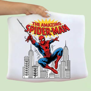 Puede incluir: Una camiseta blanca con un estampado gr&aacute;fico de Spider-Man con traje rojo y azul balance&aacute;ndose de una telara&ntilde;a. Sobre el personaje, el texto THE AMAZING SPIDER-MAN aparece en letras rojas sobre un fondo amarillo, con un horizonte urbano debajo.