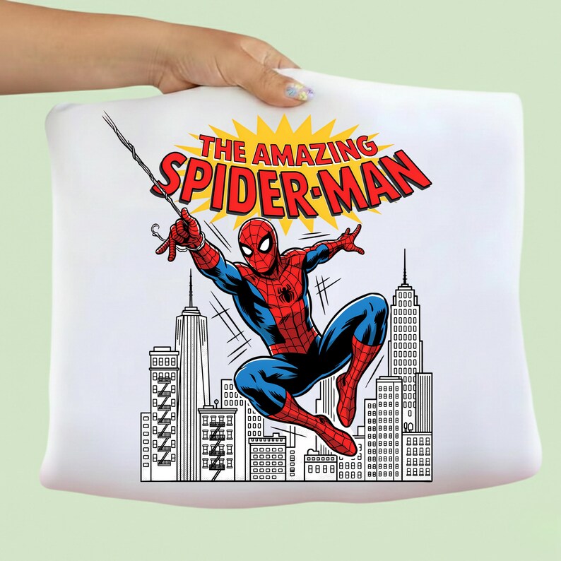 Puede incluir: Una camiseta blanca con un estampado gr&aacute;fico de Spider-Man con traje rojo y azul balance&aacute;ndose de una telara&ntilde;a. Sobre el personaje, el texto THE AMAZING SPIDER-MAN aparece en letras rojas sobre un fondo amarillo, con un horizonte urbano debajo.