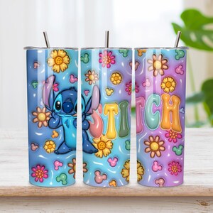 May include: A cylindrical metal tumbler with a straw, featuring a blue and purple gradient background decorated with yellow and pink flowers, small mouse-ear silhouettes, and a blue alien character next to the colorful text STITCH in bubble letters.
