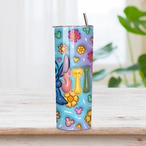 May include: A cylindrical stainless steel tumbler featuring a light blue background with a repeating pattern of colorful flowers and small mouse-ear silhouettes. The blue character Stitch is visible on the side, along with the letters STI in a puffy font.