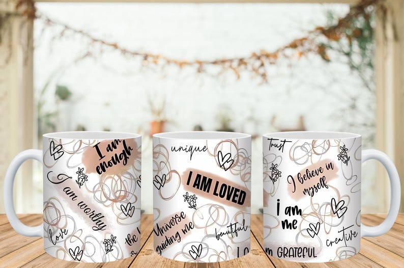 May include: White ceramic mugs with inspirational phrases like "I am enough" and "I am loved" in black script. The mugs feature abstract line art, hearts, and floral accents in a neutral color palette. The mugs are on a wooden surface.