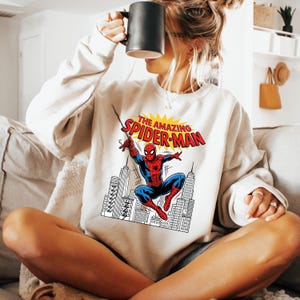 Puede incluir: Una sudadera de cuello redondo color crema con un estampado gr&aacute;fico de Spider-Man balance&aacute;ndose sobre el horizonte de una ciudad. El texto sobre el personaje dice THE AMAZING SPIDER-MAN en letras rojas y amarillas. Tiene mangas largas.