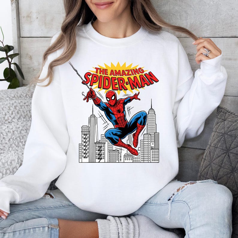 Puede incluir: Una sudadera blanca de manga larga con un estampado gr&aacute;fico de Spider-Man balance&aacute;ndose sobre una ciudad. Sobre el personaje, el texto THE AMAZING SPIDER-MAN aparece en letras rojas sobre un fondo amarillo en forma de estallido.
