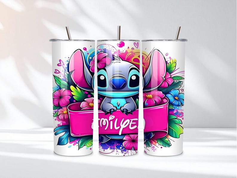K&ouml;nnte beinhalten: Ein wei&szlig;er Becher mit einer farbenfrohen Illustration von Stitch aus Lilo & Stitch. Stitch ist von rosa, blauen und lila Blumen umgeben, mit einem rosa Banner mit der Aufschrift "Emily". Der Becher hat einen silbernen Deckel und einen Strohhalm.