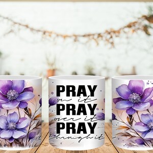 May include: White ceramic coffee mug featuring watercolor purple flowers and the text "PRAY on it, PRAY over it, PRAY through it." The floral design includes purple and lavender blooms with green stems and leaves. The mug has a white handle.