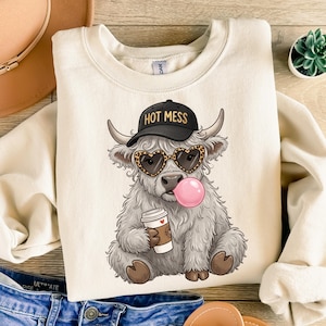 Può includere: Felpa color crema con un cartone animato di una mucca delle Highlands che indossa un berretto nero con la scritta "HOT MESS", occhiali da sole a forma di cuore con stampa leopardata e una bolla di gomma da masticare. La mucca tiene in mano una tazza di caffè.