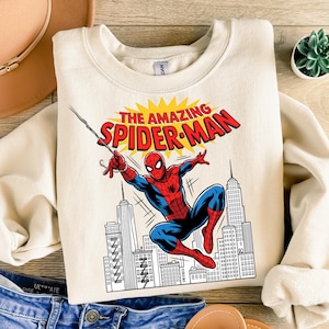 Puede incluir: Una sudadera de cuello redondo color crema con un estampado gr&aacute;fico de Spider-Man balance&aacute;ndose a trav&eacute;s de un paisaje urbano. El texto sobre el personaje dice THE AMAZING SPIDER-MAN en letras rojas sobre un fondo amarillo.