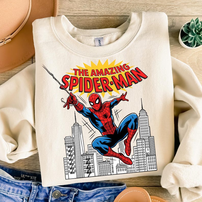 Puede incluir: Una sudadera de cuello redondo color crema con un estampado gr&aacute;fico de Spider-Man balance&aacute;ndose a trav&eacute;s de un paisaje urbano. El texto sobre el personaje dice THE AMAZING SPIDER-MAN en letras rojas sobre un fondo amarillo.