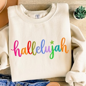 May include: A cream-colored crewneck sweatshirt featuring the word "hallelujah" printed in a vibrant, multi-colored script font across the chest. Each letter is a different color, and a small green star sits above the letter "j".