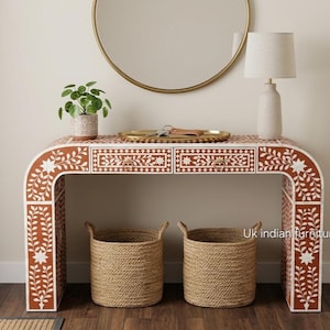 May include: A rectangular console table with a curved frame featuring a terracotta and white floral inlay pattern. The table has two drawers with small circular knobs and is positioned above two woven natural fiber baskets on the floor.