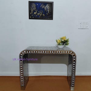 May include: A decorative table with a curved design, featuring a brown and white inlaid pattern. The table has a rectangular top and is adorned with a diamond pattern along the edges. A framed artwork with deer and trees hangs above the table.