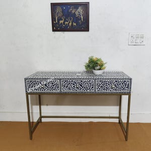 May include: A decorative desk with a navy blue and white floral pattern on the drawers and tabletop. The desk has three drawers with gold-colored knobs and a gold-colored metal frame. A small plant in a white pot sits on top of the desk.