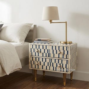 May include: A two-drawer nightstand featuring a cream and navy blue geometric inlay pattern, four tapered wooden legs with gold-toned metal caps, and round knobs. A brass-finished desk lamp with a beige fabric shade sits on the top surface.