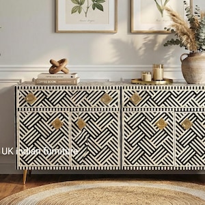 May include: A black and white geometric patterned cabinet with gold handles. The cabinet has four doors and sits on gold legs. A potted plant and decorative items are on the left. A vase of dried flowers sits on top.